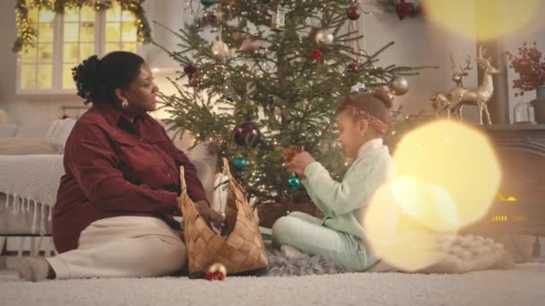 Linssi leimahti laukaus onnellinen Afrikkalainen amerikkalainen äiti ja pieni tytär chattailuun samalla koristelu joulukuusi yhdessä viihtyisässä asunnossaan - Materiaali, video