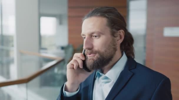 Medium close up of businessman talking to client on mobile phone while walking in office hall - Footage, Video