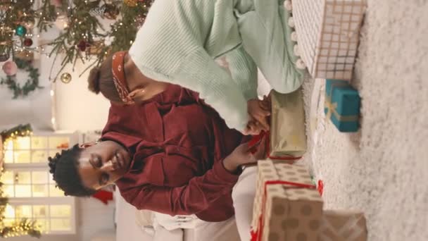 tiro vertical de juguetona niña afroamericana ayudando a mamá a atar lazo en regalo de Navidad con cinta roja, sentados juntos en el suelo en acogedor apartamento decorado - Imágenes, Vídeo