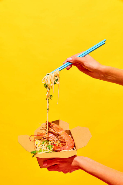 Неузнаваемая женщина ест вкусные горячие ВОК с креветками с палочкой для еды из бумажной коробки изолированный желтый студийный фон Концепция национальной кухни, кухни, ресторанного меню, питания, доставки. Объявление - Фото, изображение