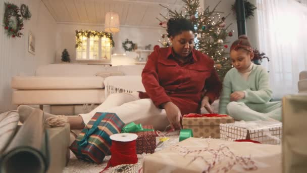 Espectáculo de puñalada de ángulo bajo de mamá e hija afroamericanas sentadas en el suelo en una acogedora sala de estar decorada para envolver navidades y obsequiar regalos en cajas - Imágenes, Vídeo