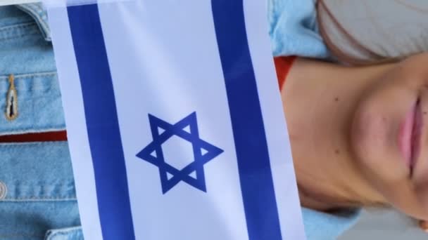 Belle adolescente caucasienne en t-shirt rouge et veste en denim gardant petit drapeau d'Israël dans ses mains. Une jeune femme se tient debout à l'extérieur, souriante et agitant un petit drapeau israélien dans ses mains. Réel - Séquence, vidéo