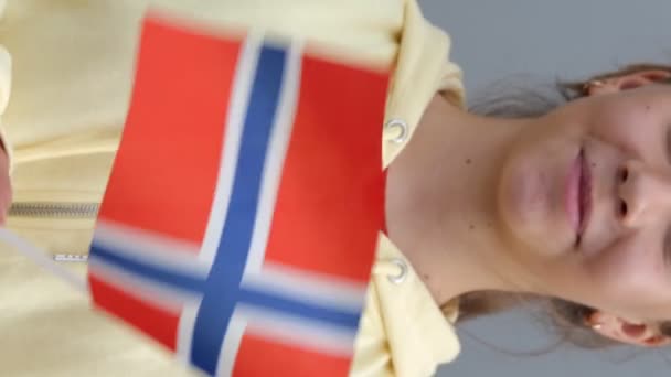 Caucasico giovane bella adolescente in felpa gialla con cappuccio sventola piccola bandiera norvegese, sorridendo alla macchina fotografica. Zoom out, primo piano, sfondo grigio, una piccola femmina tiene la bandiera della Norvegia nelle sue mani - Filmati, video