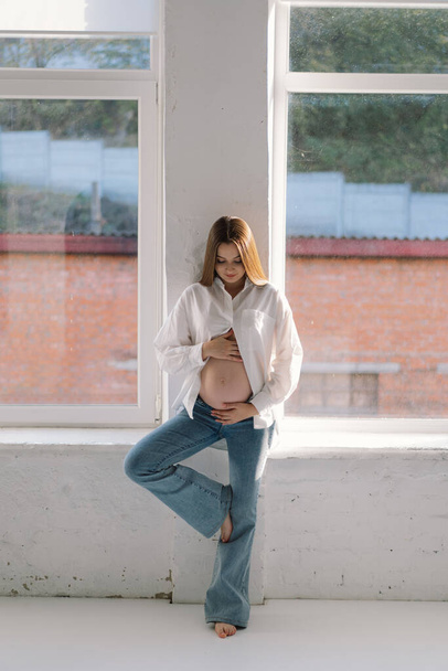Мила молода дівчина сидить на білій студії в теплому світлі з руками на вагітному животі на стіні з тіні. Вагітна концепція, вільний простір, насолоджується вагітністю. Вагітне тіло. - Фото, зображення