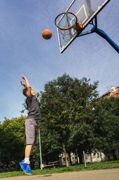 Mignon garçon en t-shirt gris joue au basket sur l'aire de jeux de la ville. - Photo, image