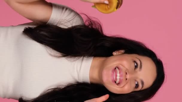 Uśmiechnięta kobieta bawiąca się podczas tańca i trzymając hamburgera śpiewającego piosenkę, odizolowana na różowym tle. Ludzie, styl życia, koncepcja śmieciowego jedzenia. Pionowe wideo w czasie rzeczywistym - Materiał filmowy, wideo