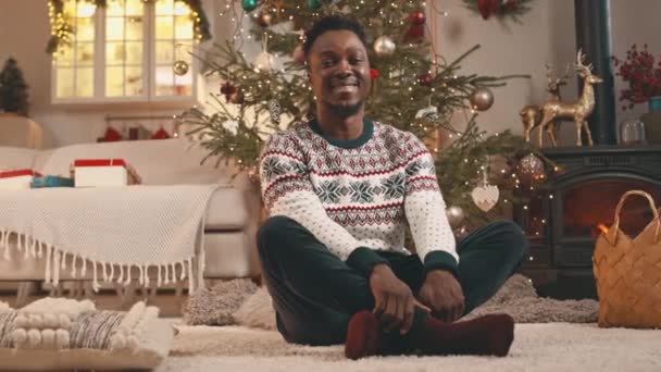 Portrét nadšené mladé Afroameričan muž na sobě bílé vánoční svetr sedí na podlaze krásně zdobené vánoční stromeček v útulném studio apartmánu - Záběry, video