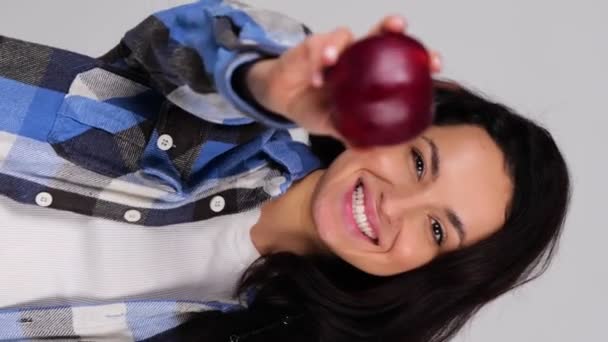 Hembra sonriente mostrando manzana roja mientras la muerde, aislada sobre fondo gris, espacio para copiar. Concepto de comida sana y fresca. Vídeo vertical en tiempo real - Imágenes, Vídeo