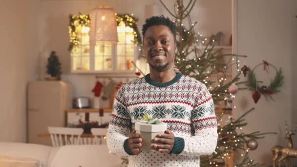 Retrato medio del joven alegre hombre negro en jersey de Navidad posando para la cámara con caja de regalo de Navidad en las manos de pie en acogedor apartamento estudio decorado para Navidad - Imágenes, Vídeo
