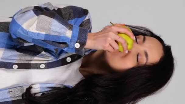ジューシーな黄色のりんごを噛んだ平らなシャツで美しい女性を笑顔にします. 灰色の背景に立っている女性. 健康で新鮮な食品コンセプト。 リアルタイムの垂直ビデオ - 映像、動画