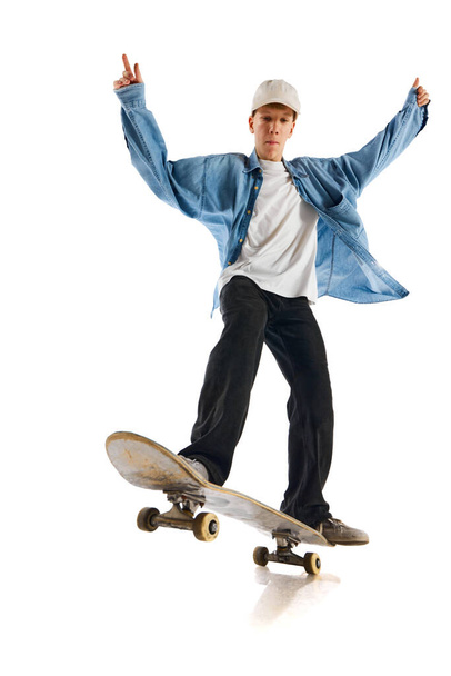 Teen boy in blue jeans shirt and cap in motion, training with skateboard, doing stunts isolated over white background. Concept of professional sport, competition, training, action. Copy space for ad - Foto, Imagem