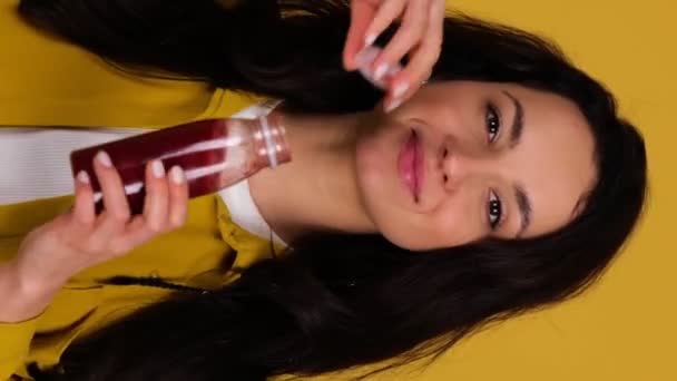 Mosolygó fiatal nő élvezi inni a friss gyümölcslevet, miközben pózol a stúdióban. Méregtelenítő diéta. Táplálkozás egészséges koncepció. Valós idejű függőleges videó - Felvétel, videó