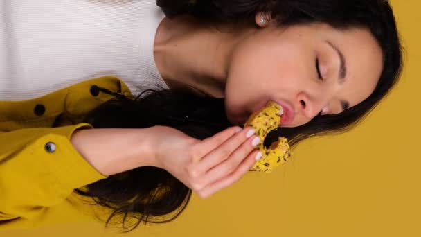 Mulher feliz desfrutando de um donut enquanto morde. Senhora de pé contra fundo amarelo. Um conceito saudável de comida de plástico. Vídeo vertical em tempo real - Filmagem, Vídeo