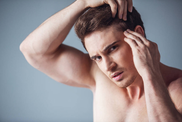 Handsome young man with bare torso is combing hair and looking at camera, on gray background - Photo, Image