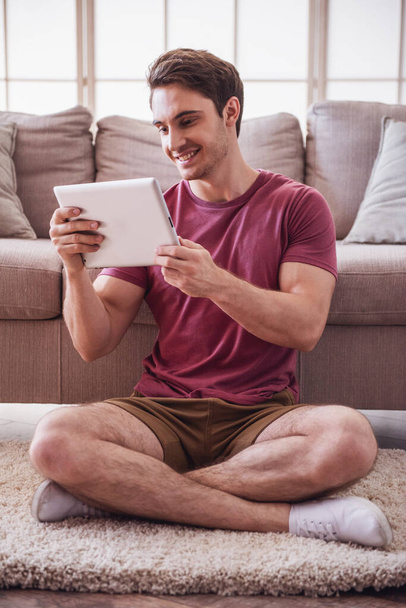 Όμορφος άντρας σε casual ρούχα είναι χρησιμοποιώντας ένα ψηφιακό tablet και χαμογελώντας ενώ κάθεται στο πάτωμα στο σπίτι - Φωτογραφία, εικόνα
