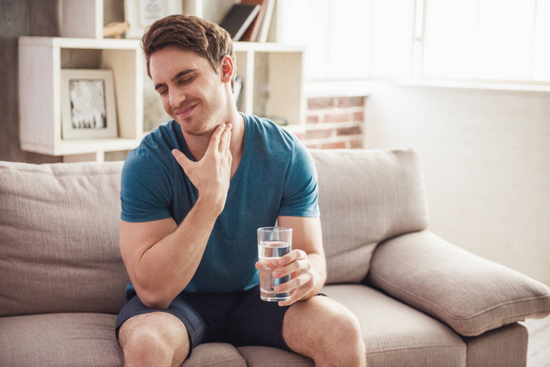 Красивый парень в повседневной одежде держит стакан воды и трогает его за горло, сидя дома на диване
 - Фото, изображение