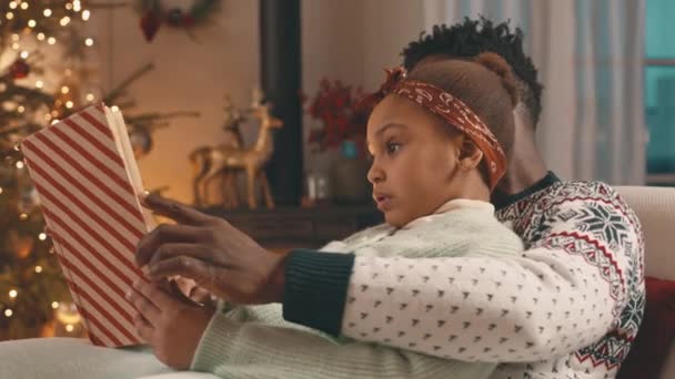 Chest up of African American man with little daughter reading book together sitting on couch by fireplace in cozy living room decorated for Christmas - Footage, Video