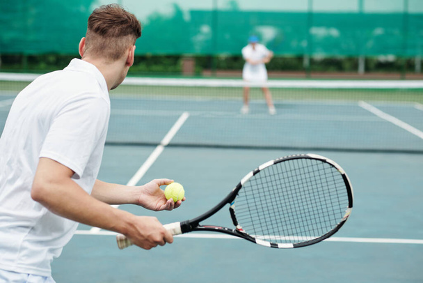 Young man playing tennis with friend and serving ball, view from back - Foto, afbeelding