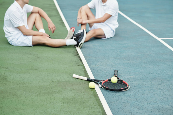 Tennis rackets and balls on court next to sportsmen resting after game - Photo, Image