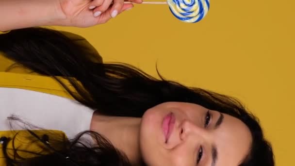 Lachende jonge vrolijke vrouw met lolly en likken, terwijl ze poseert in de studio, geïsoleerd op gele achtergrond. Real-time verticale video - Video