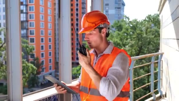Mies rakentaja kypärä kanssa radiopuhelin tilalla digitaalinen tabletti työskennellessään ulkona katolla. Liiketoiminta, rakentaminen, teollisuuden käsite. Reaaliaikainen - Materiaali, video