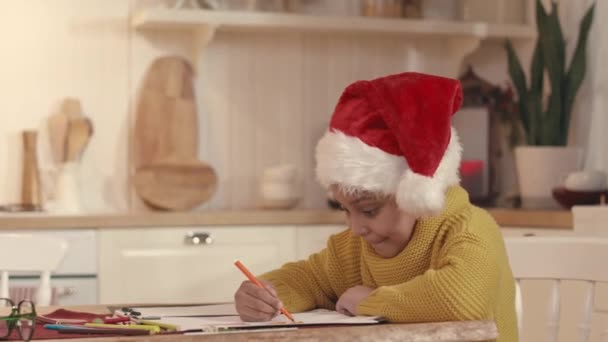 Chest up of cute African American little girl wearing red Santa hat and mustard sweater drawing while sitting at kitchen table in cozy apartment decorated for Xmas - Footage, Video