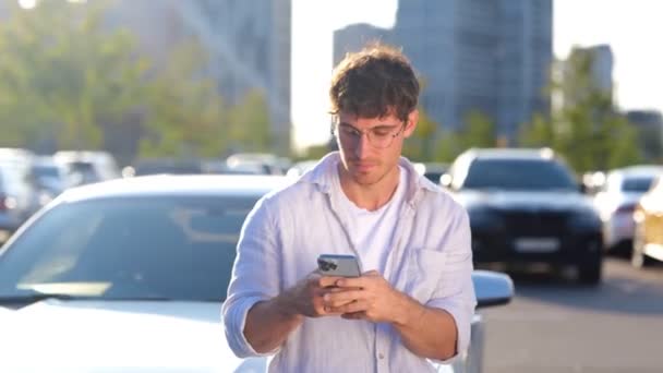 Щасливий молодий чоловік стоїть біля своєї машини, тримаючи смартфон, друкуючи повідомлення. Транспорт, технології та концепція способу життя. Реальний час - Кадри, відео
