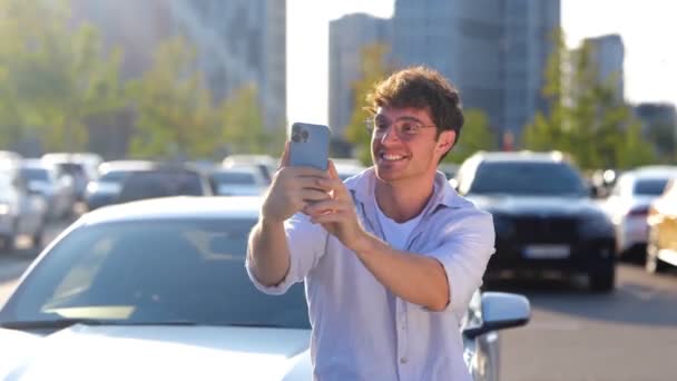 Glimlachende man die een smartphone vasthoudt en buiten foto 's maakt van zijn nieuwe auto in de stad. Vervoer, technologie en lifestyle concept. Real time - Video