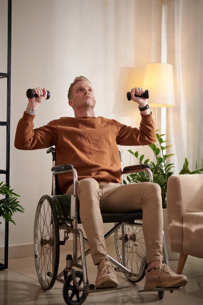 Jovem com deficiência sentado em cadeira de rodas e fazendo exercícios com pequenos halteres - Foto, Imagem