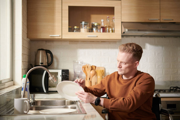 Vakava nuori mies pyörätuolissa tiskaamassa astioita päivällisen jälkeen - Valokuva, kuva