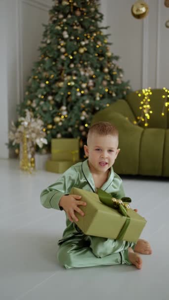 Porträt eines fröhlichen kaukasischen Jungen, der versucht ist zu erraten, welches Geschenk sich in der glänzenden Weihnachtsgeschenkschachtel befindet. Überraschte glückliche Gesichter eines Kindes mit einem Weihnachtsgeschenk - Filmmaterial, Video