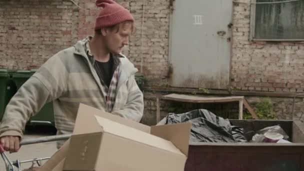 Inclinazione verso il basso colpo di senzatetto rovistando in bidoni della spazzatura in cerca di vestiti sulla strada - Filmati, video