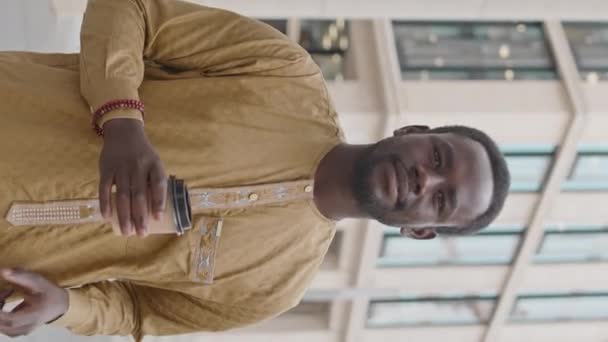 Retrato vertical mediano del joven afroamericano encantado con camisa amarilla tradicional sonriendo a la cámara, parado al aire libre con una taza de café de papel en la mano - Imágenes, Vídeo