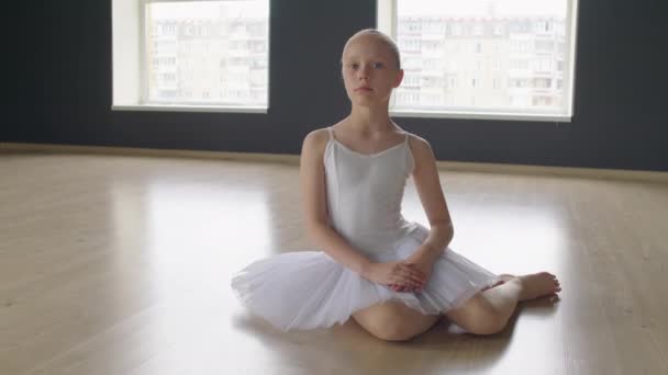 Portré felvétel kis balett iskolás táncosról, aki a stúdió padlóján ül és a kamerát nézi. - Felvétel, videó