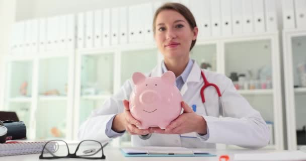 Médico terapeuta con alcancía rosa en las manos en la clínica. Procedimientos médicos costosos e inversiones en costos de seguro de salud y salud - Metraje, vídeo