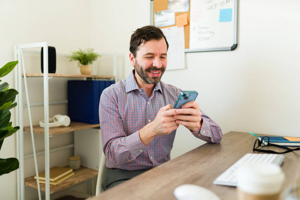 Latynoski szczęśliwy przedsiębiorca lub prawnik uśmiechnięty podczas pisania SMS-ów na smartfonie podczas pracy w biurze - Zdjęcie, obraz