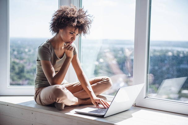 Kaunis tyttö työskentelee kannettavan tietokoneen kanssa istuessaan ikkunalaudalla kotona - Valokuva, kuva