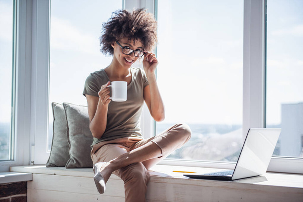 A gyönyörű üzletasszony laptopot használ, kávét iszik és mosolyog, miközben otthon ül az ablakpárkányon. - Fotó, kép