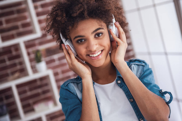 Hermosa chica en los auriculares está escuchando música, mirando a la cámara y sonriendo mientras pasa tiempo en casa - Foto, imagen