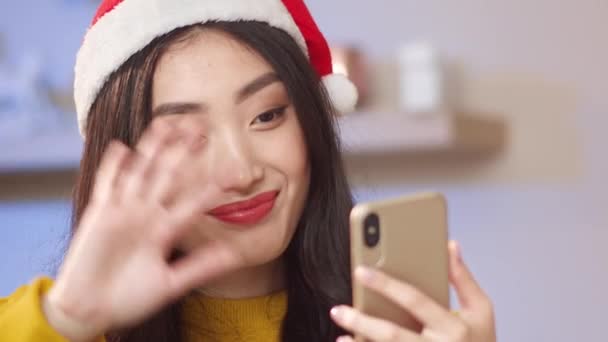 Щаслива азіатська дівчина з різдвяним капелюхом махає рукою на камеру телефону під час віддаленого зв'язку. Симпатична жінка говорить на смартфоні слова привітання з зимовими канікулами своїм друзям або родині. - Кадри, відео