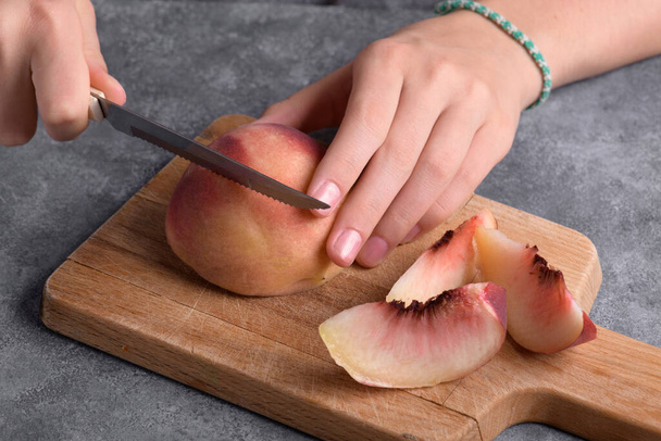 Authentic female hands cutting a fresh ripe peach on wooden cutting board on a grey table background, close up. Healthy summer snack, food lifestyle - Photo, image