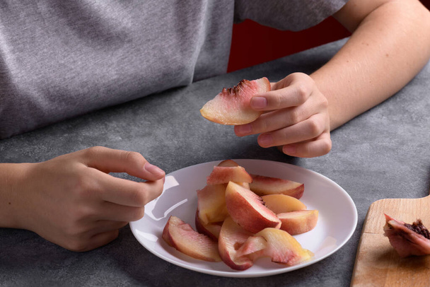 Authentic female hands holding a piece of a fresh cutting ripe peach on white plate on a grey table background, close up. Healthy summer snack, food lifestyle - Foto, imagen