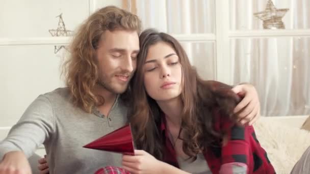 Любимая кавказская пара проводит время на диване в уютной квартире. Заботливый молодой парень обнимает свою милую девушку, которая держит бумажную шляпу для празднования. У людей романтический вечер. Концепция отношений. - Кадры, видео