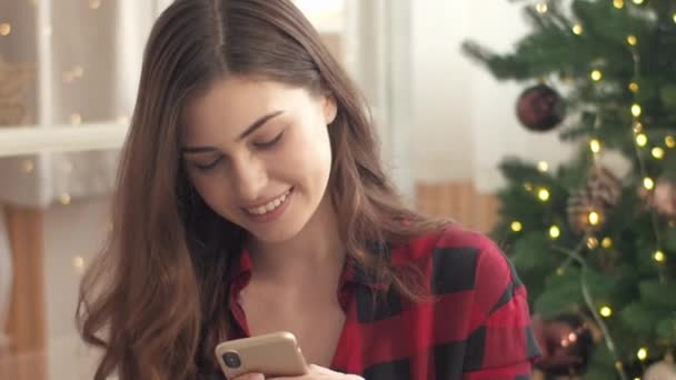 Mooi blank meisje met een geruit shirt dat online communiceert met haar mobiele telefoon. Actief typen bericht op de telefoon aan haar vrienden tijdens het zitten in de buurt van de kerstboom met slinger. Technologie gebruiken. - Video