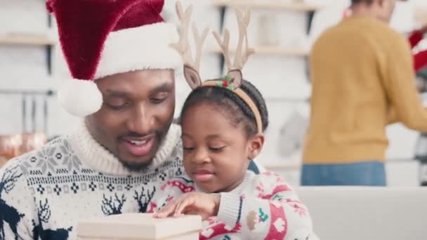 Szczęśliwej afroamerykańskiej rodziny obchodzącej święta w domu. Tata w kapeluszu Świętego Mikołaja daje prezent przytulając swoje kochające dziecko. Pudełko dziecięce i miękka zabawka. Wesołych świąt. - Materiał filmowy, wideo