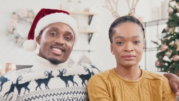 Retrato de casal afro-americano vestindo camisolas de malha sentado no sofá. Homem atraente com chapéu de Papai Noel abraçando sua esposa bonita que na cabeça de chifres de veados. Espírito de Natal. - Filmagem, Vídeo
