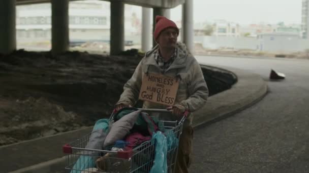 Medium lange schot van dakloze man in lompen kijken rond en kiezen richting gaan onder de brug met winkelwagentje in de snelweg gebied - Video