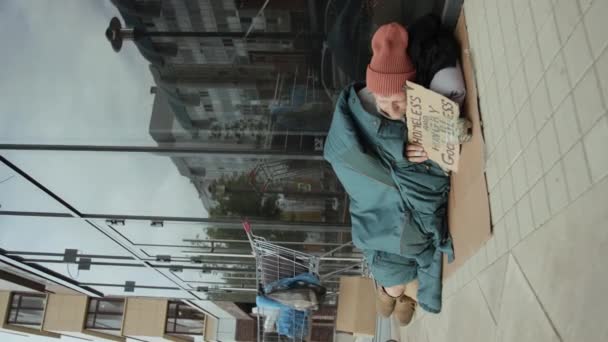 Colpo verticale di senzatetto con cartello che dorme su cartone che dorme accanto al carrello della spesa con la sua roba vicino all'edificio - Filmati, video