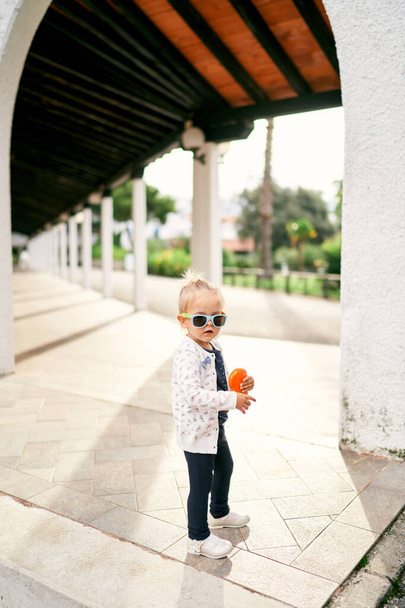 Маленька дівчинка з іграшкою в руках стоїть в довгій перголі з колонами в саду. Високоякісна фотографія - Фото, зображення