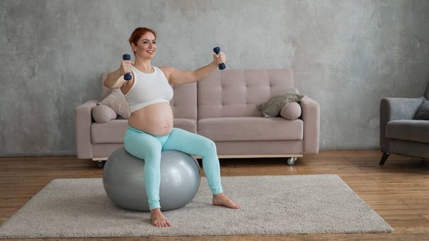Schwangere macht Übungen mit Kurzhanteln, während sie zu Hause auf einem Fitnessball sitzt - Foto, Bild